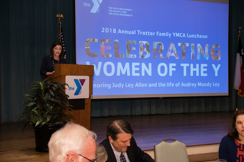 A woman dressed in black stands at a YMCA podium with a projector screen behind her that reads "Celebrating Women of the Y"