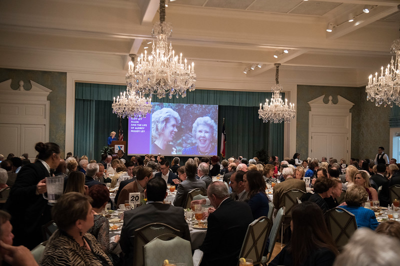 Over a crowd of people sitting at round tables in a formal dining hall, a projector screen shows Judy Ley Allen and Audrey Moody Ley as Judy stands at a podium and addresses the audience.