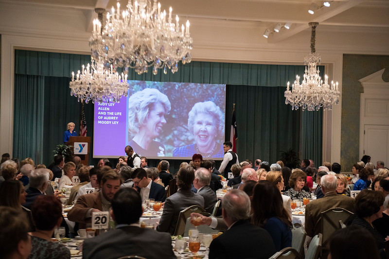 Over a crowd of people sitting at round tables in a formal dining hall, a projector screen shows Judy Ley Allen and Audrey Moody Ley as Judy stands at a podium and addresses the audience.
