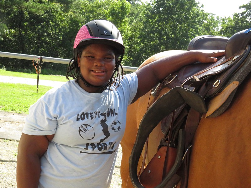 A young girl wears a horse riding helmet as she stands with her arm up on the horse saddle of the mare next to her