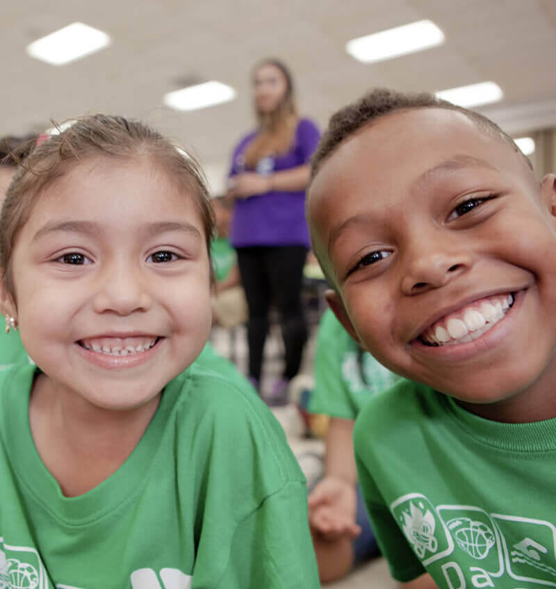 Two children in green shirts smile into the camera at summer camp. On the left, a young girl with her dark hair back in a ponytail next to a boy of darker complexion and a buzz cut to the right.