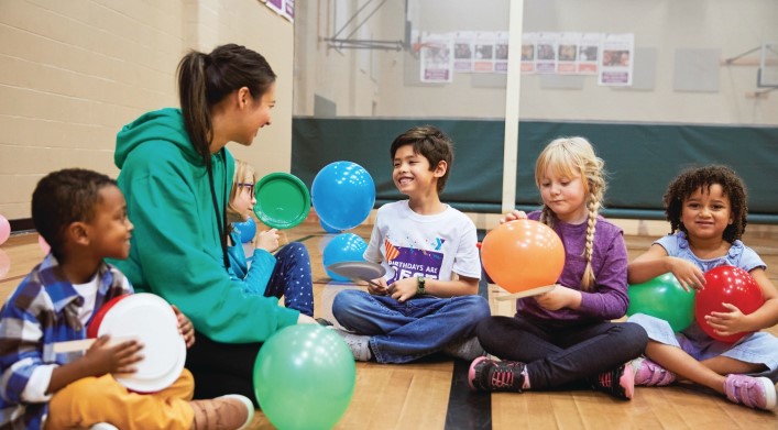 A group of children and an adult sitting in a circle in a gymnasium while holding balloons