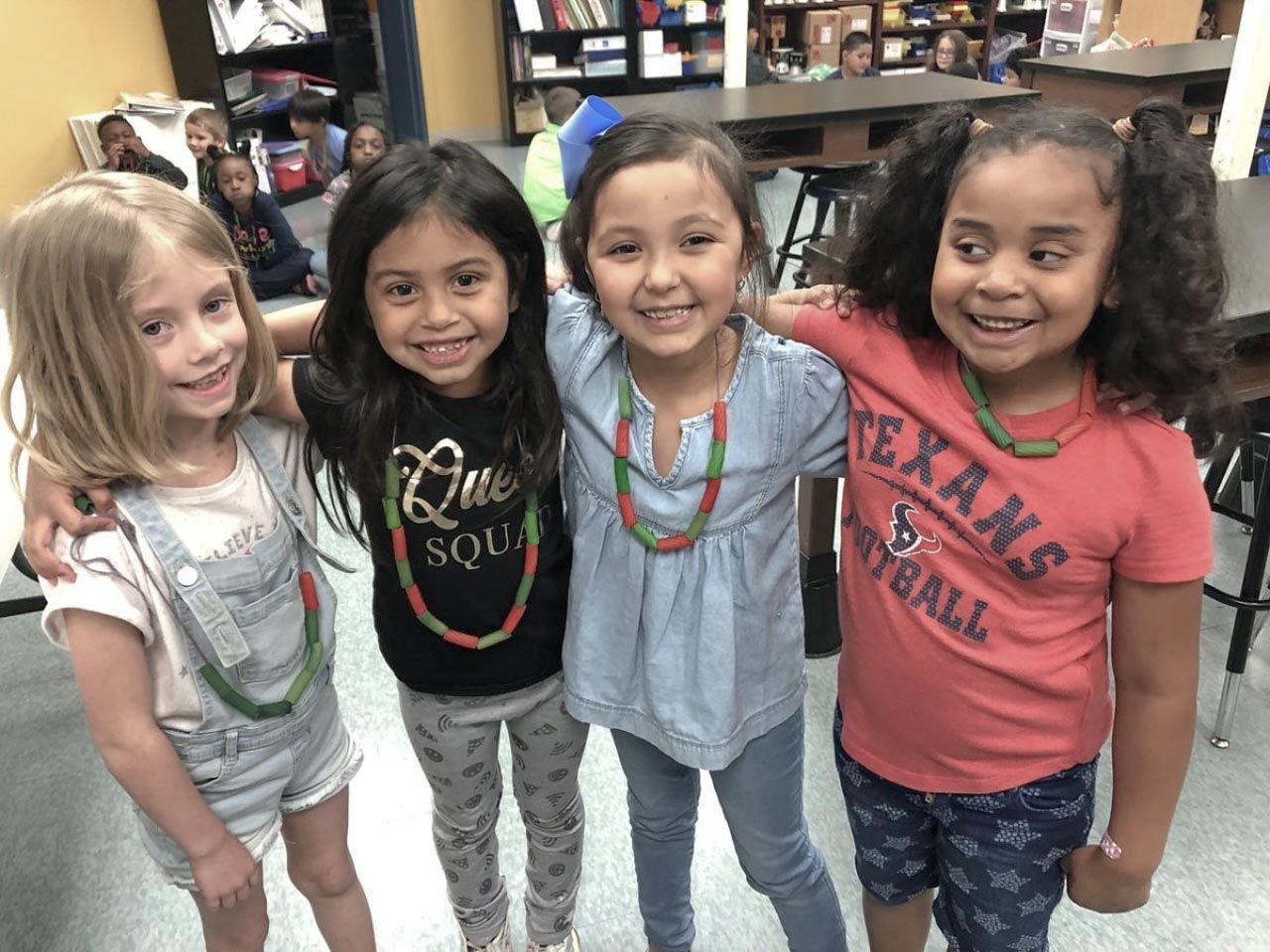 Four young girls stand together with their arms over each other's shoulders in a line. The two girls on the left have their hair down, while the third has her dark hair in a ponytail, and the front has in curly pigtails. All four girls are smiling at the camera during afterschool care.