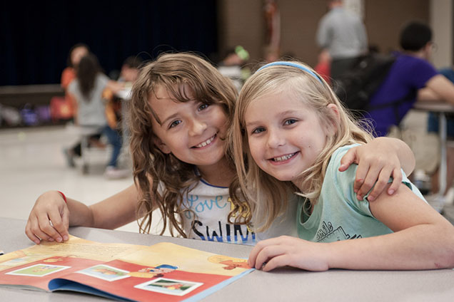 Two girls sit side-by-side, smiling with their hair down as the girl on the left has her arm over the shoulder of the girl on the right as they look into the camera at afterschool care while reading a book.