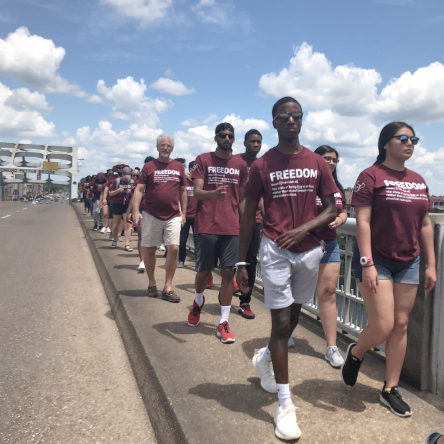 A group of teens in matching maroon shirts walks on the sidewalk outside
