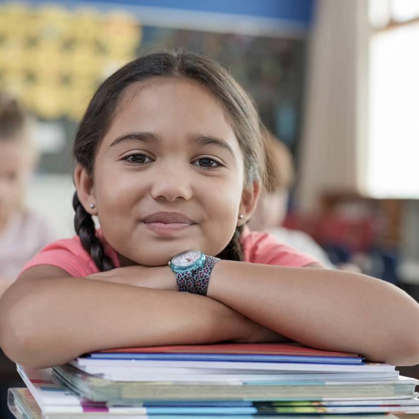 Hispanic girl with chin on books Portrait of cute little schoolgirl leaning on stacked books in classroom. Happy young latin girl in casual keeping chin on notebooks. Closeup face of smiling girl at elementary school.