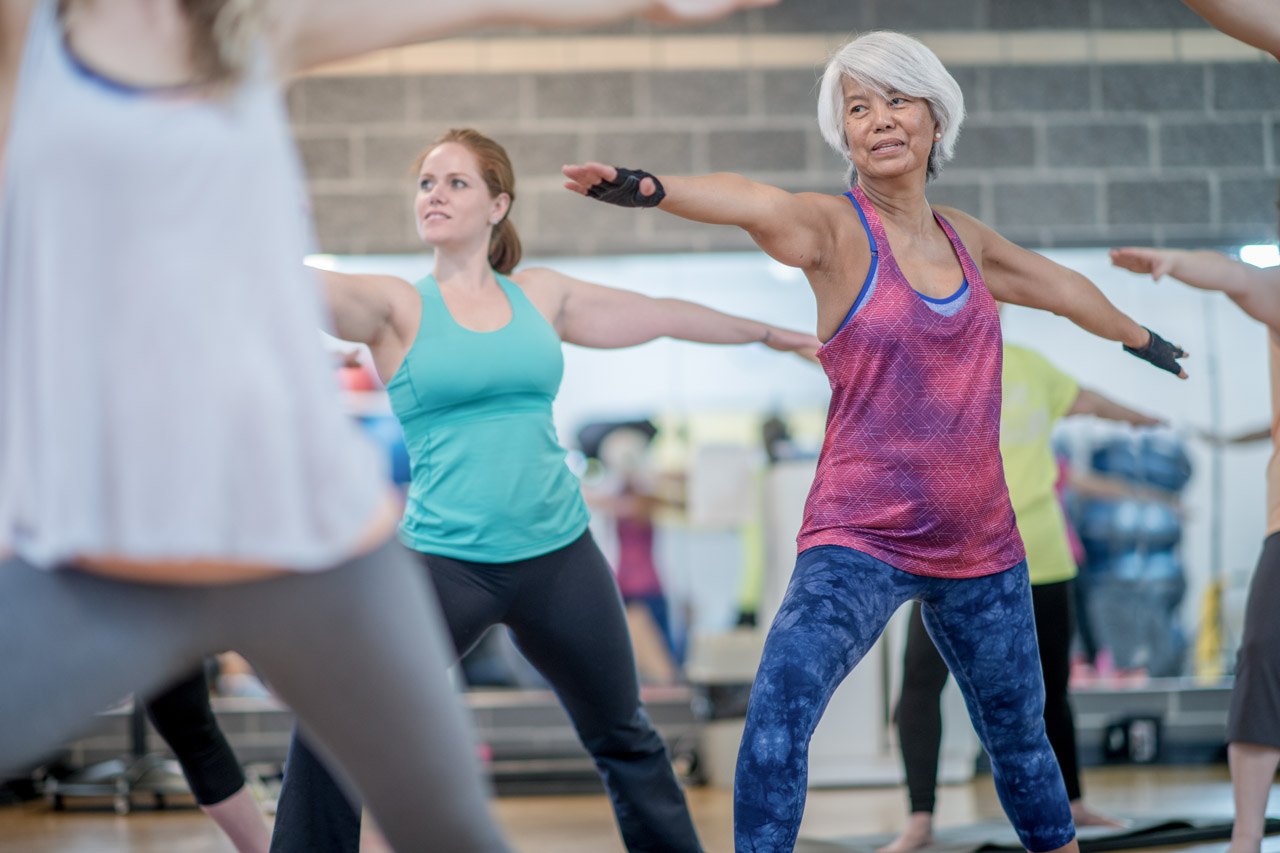 A group of adults are working out together at a fitness center. Two women are doing yoga while standing. They have their arms outstretched.