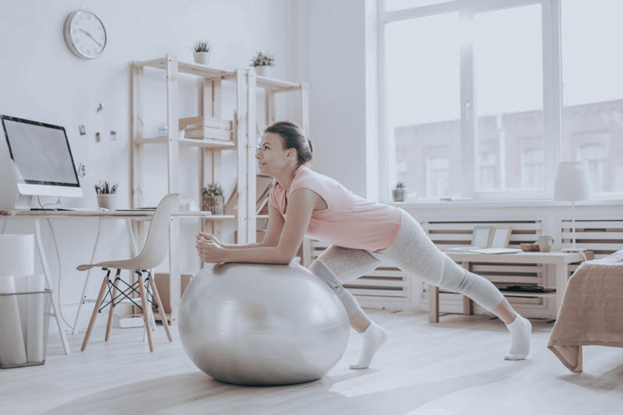 Full length portrait of beautiful mixed race woman exercising on fitness ball during home workout in sunlight, copy space