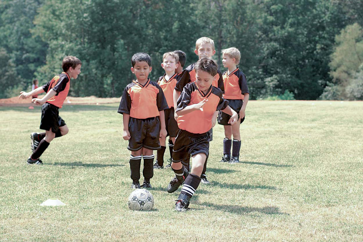 A group of elementary aged children play soccer in orange and black uniforms