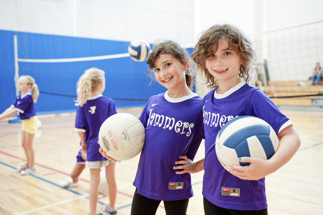 Two children in purple jerseys holding volleyballs in a gymnasium.