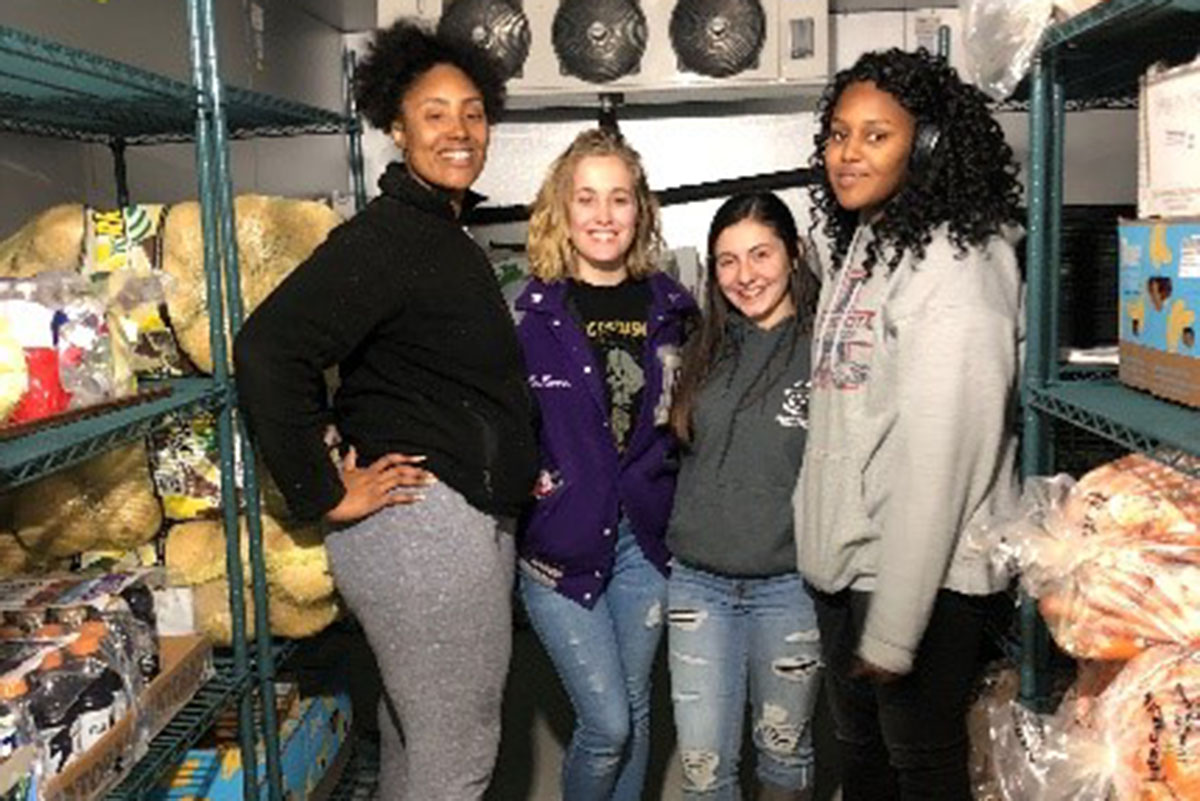 Four people standing in a storage room with shelves of food items.