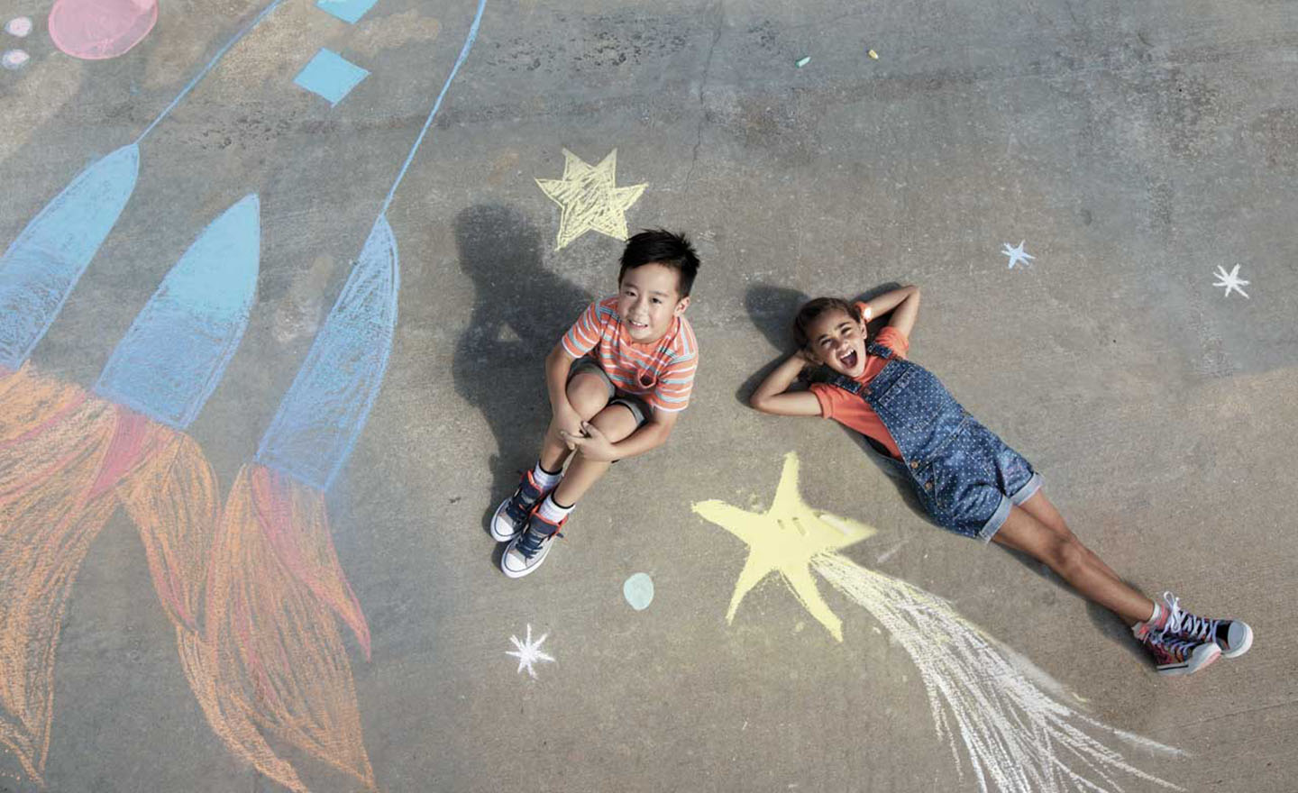 Two children laying on a sidewalk with chalk drawings around them.