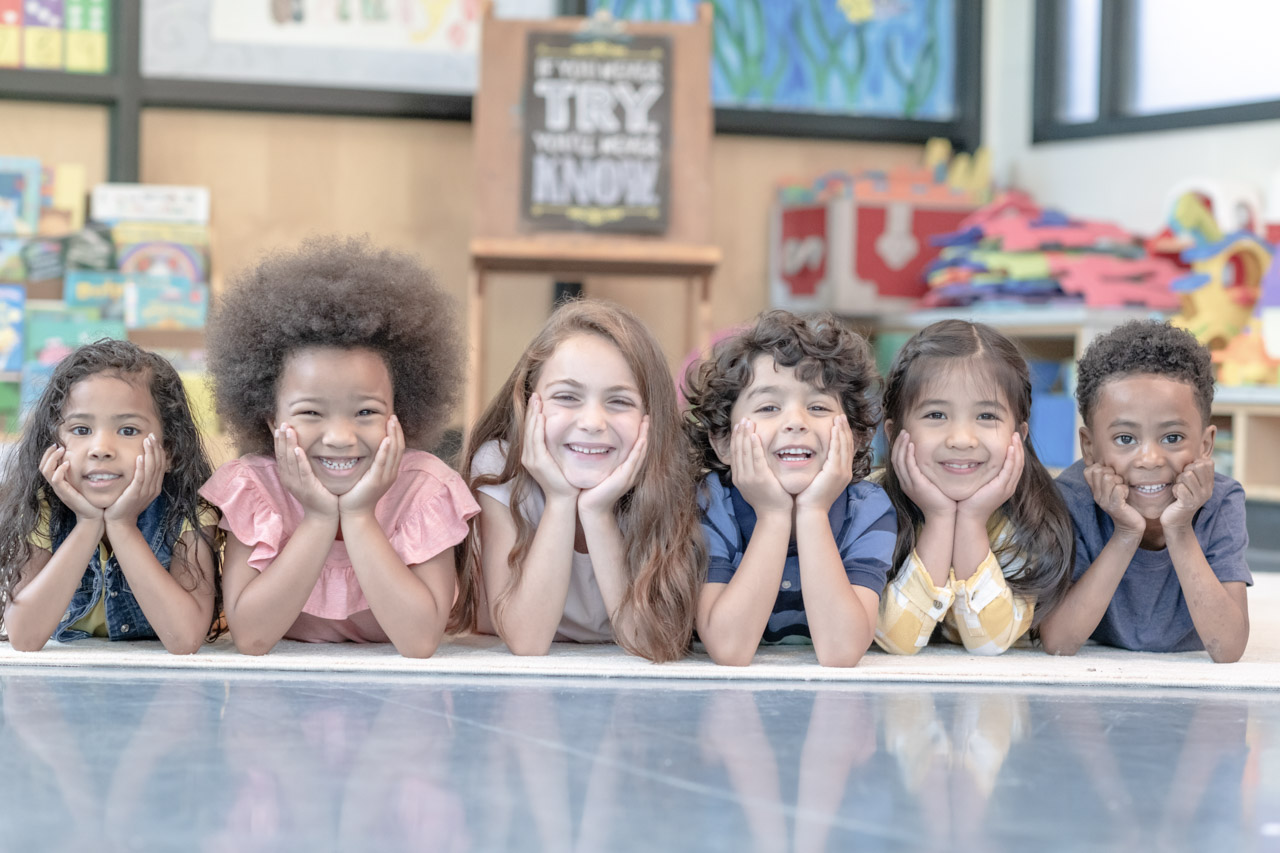 Portrait of a happy multi-ethnic group of preschool students. The cute children are laying on their stomachs in a line. They are in their classroom. The kids are resting their chins in their hands while smiling directly at the camera.