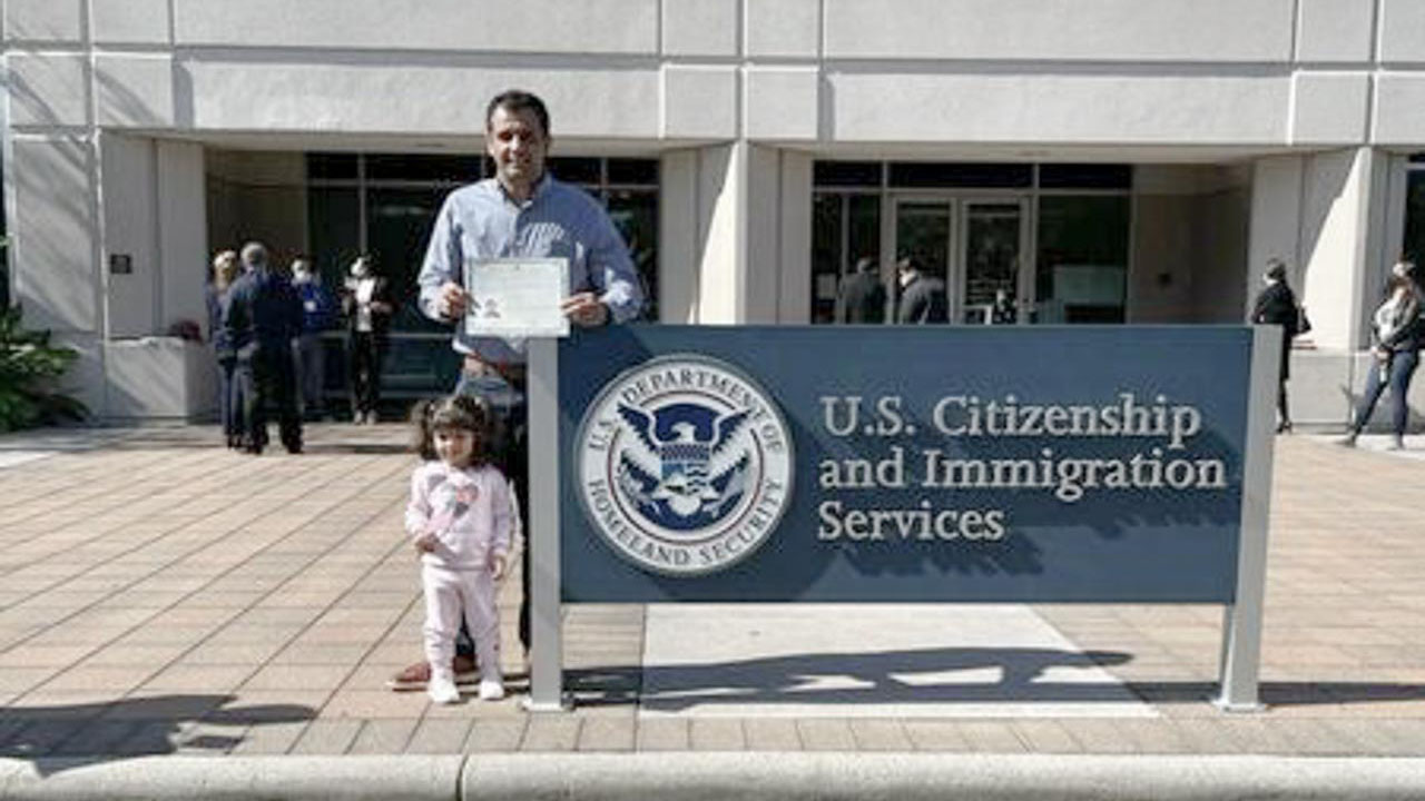 A photo of a person holding a certificate and standing next to a child in front of a U.S. Citizenship and Immigration Services sign.