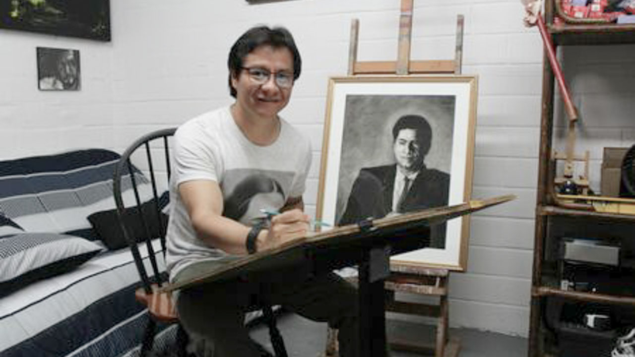 A person sitting at a desk while drawing with a sketch of a person with a blurred face in the background.