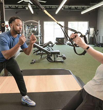 A man in a blue shirt, short dark hair, and a well-groomed beard holds up fists as he demonstrates fitness techniques to a person using tension equipment.
