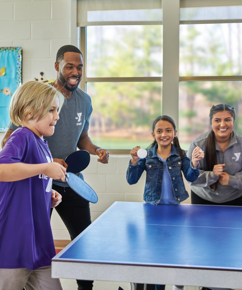 A child in a purple shirt plays pinball with a YMCA mentor while another mentor and student cheer them on.