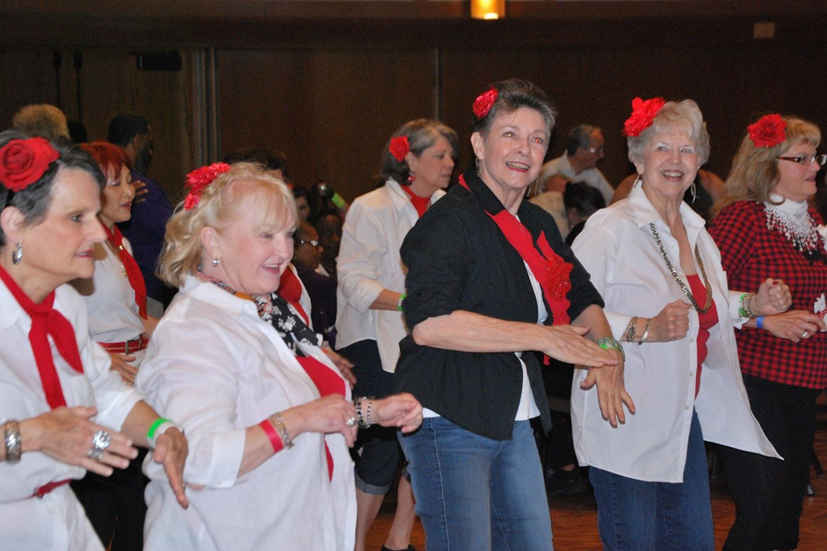 A lively group of elderly ladies dancing, wearing red scarves and flowers in their hair