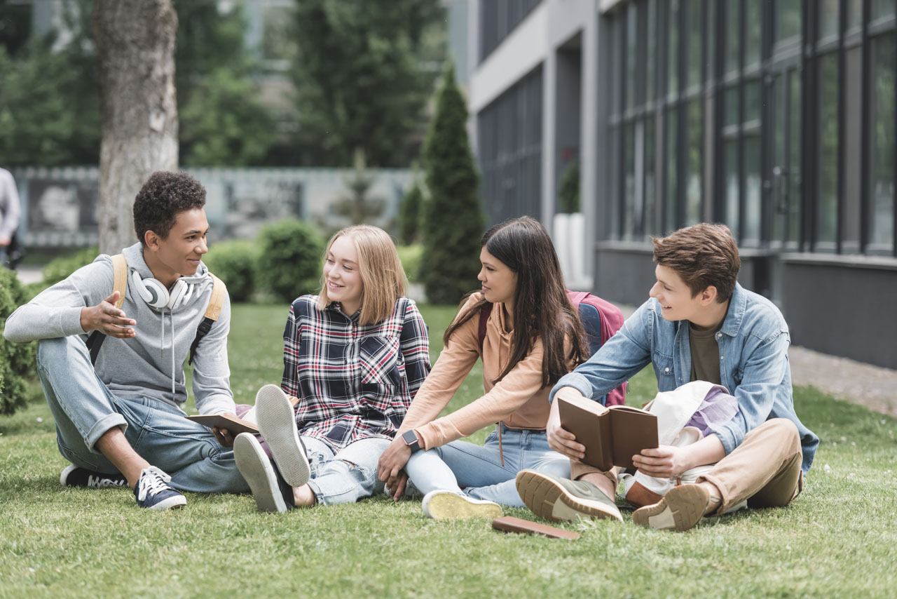 A group of four young people sitting on the grass in front of a building, reading and talking