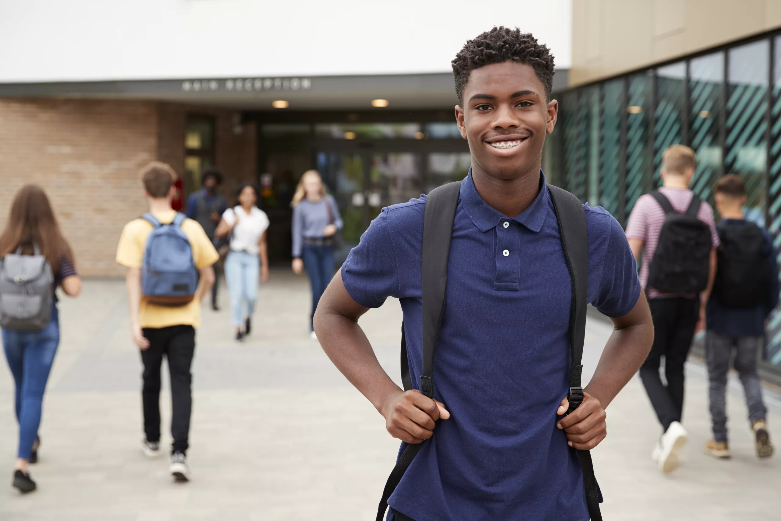 Student wearing a blue collared shirt posing with backpack, fellow students walking behind